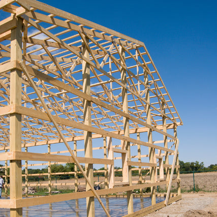 Pole BarnsWooden frame of a barn under construction against a clear blue sky, set in a rural landscape. Workers are visible in the background, suggesting progress.