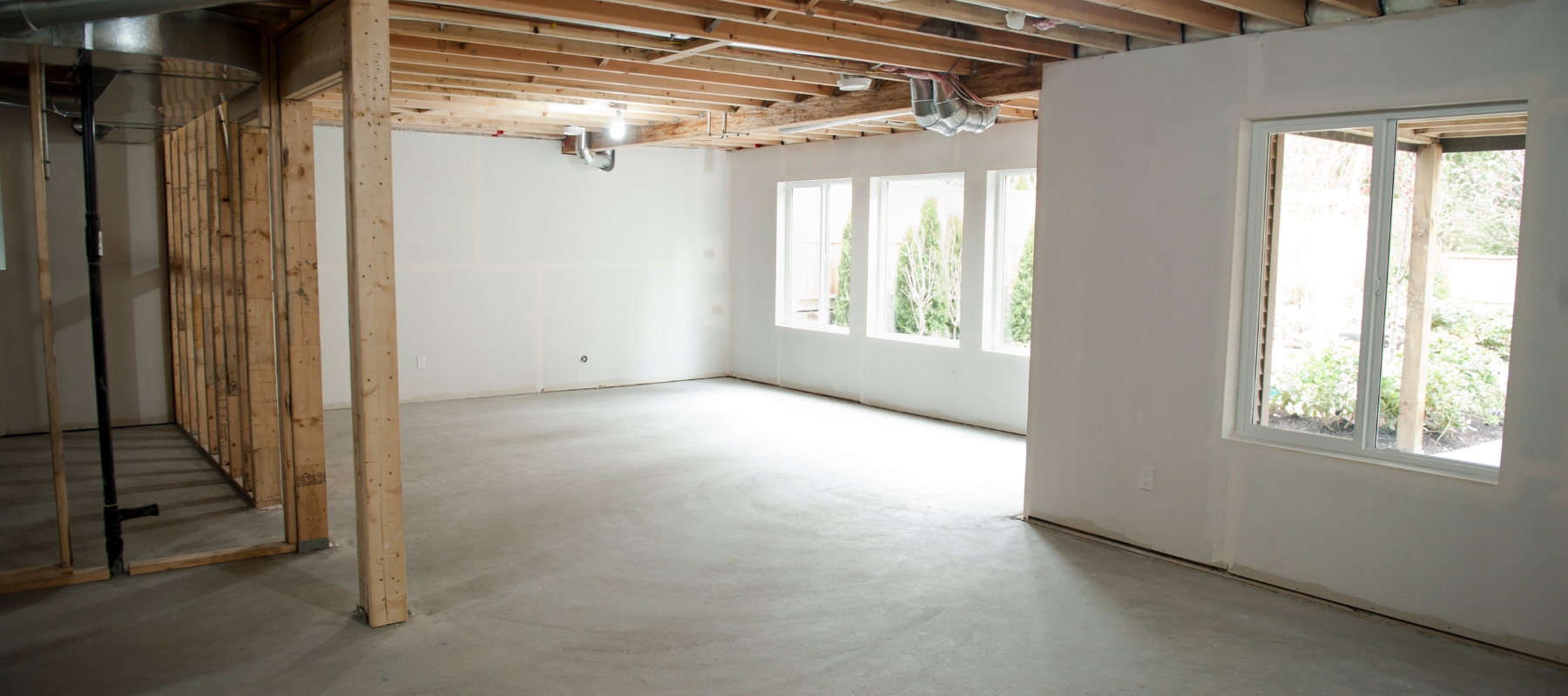 Unfinished basement with concrete floor, exposed wooden beams, and white walls. Sunlight filters through three windows, creating a bright atmosphere.
