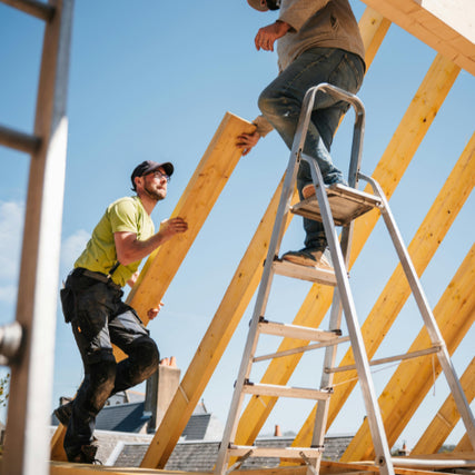 Lumber & Building MaterialsTwo construction workers assemble a wooden roof frame. One stands on a ladder, the other hands him a beam. The scene conveys teamwork under a clear blue sky.